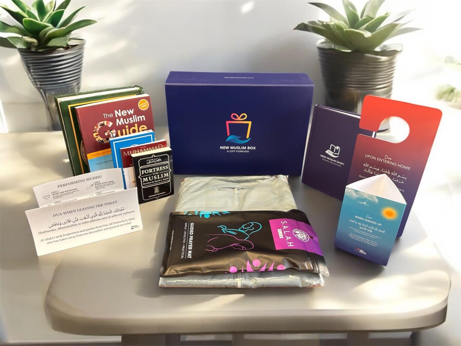 The New Muslim Box laid out: a translated Qur'an, the New Muslim Guide, Fortress of the Muslim, a guided prayer mat, prayer clothing, Zamzam water, du'a cards and door hangers, arranged on a bright table between two plants.